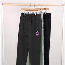 Load image into Gallery viewer, Be Happy black joggers from the Pink Runway Bright Future range hanging on a rail with 2 other pairs of jogging bottoms. There is a drippy smiley face in pink/purple ombre under the left pocket.